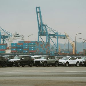 A lineup of parked cars at a bustling industrial shipping port with cranes and containers.