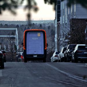 Amazon delivery van driving through an urban street at twilight with cars parked along the side.